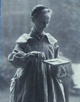 Mary Watts in a long dress and apron, holding a small palette, looking down with a focused expression.