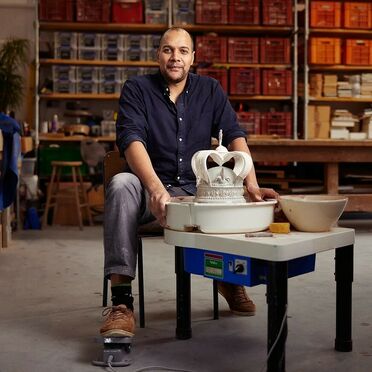 A man, seated in a pottery studio, smiling at the camera, with a finished white ceramic crown on a pottery wheel in front of him. He wears a navy blue shirt, gray pants, and brown shoes with green and black socks. The studio has shelves of storage bins and pottery supplies in the background.