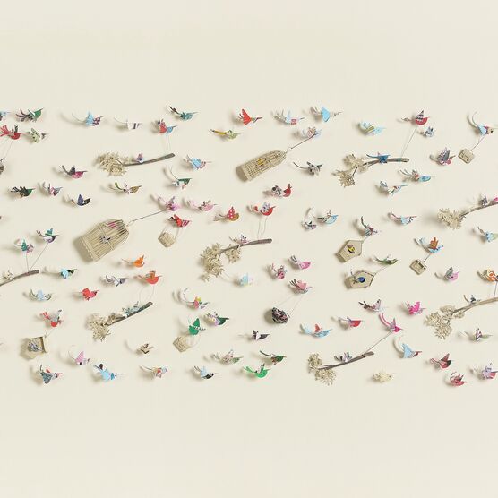 A flat, rectangular artwork against a light beige background, featuring numerous small, colourful paper birds in flight. Some birds are perched on thin branches with delicate white clusters, while others are suspended mid-air with wire-like attachments. Scattered amongst the birds are small birdhouses and cages. The title, "Crowded Skies," by Lucy Dorothy