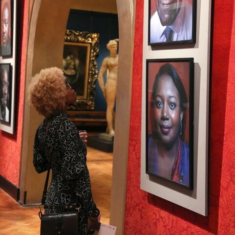 Woman with blonde afro wearing glasses, black patterned dress, and purse, looking up at portrait in a gallery. The gallery features portraits of individuals, and other art pieces including a statue and a painting on the wall.