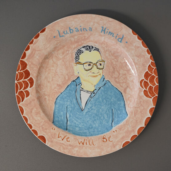 A ceramic plate decorated with a painting of Lubaina Himid. The plate is resting on a grey background. The plate is pale orange with circles of darker orange around the edge. The portrait of Himid sits in the centre of the plate, depicting a women looking off to the side, she is wearing a light blue long sleeve top and orange glasses. Above the portrait it reads 'Lubaina Himid' and below at the bottom of the plate is says 'We will be'.
