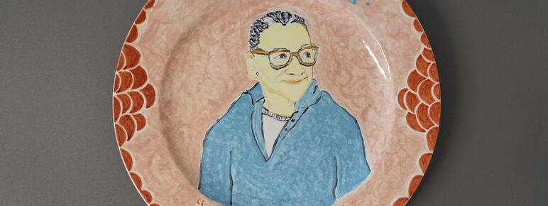 A ceramic plate decorated with a painting of Lubaina Himid. The plate is resting on a grey background. The plate is pale orange with circles of darker orange around the edge. The portrait of Himid sits in the centre of the plate, depicting a women looking off to the side, she is wearing a light blue long sleeve top and orange glasses. Above the portrait it reads 'Lubaina Himid' and below at the bottom of the plate is says 'We will be'.