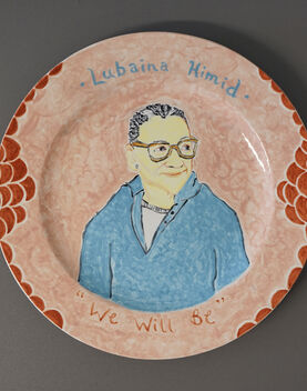 A ceramic plate decorated with a painting of Lubaina Himid. The plate is resting on a grey background. The plate is pale orange with circles of darker orange around the edge. The portrait of Himid sits in the centre of the plate, depicting a women looking off to the side, she is wearing a light blue long sleeve top and orange glasses. Above the portrait it reads 'Lubaina Himid' and below at the bottom of the plate is says 'We will be'.