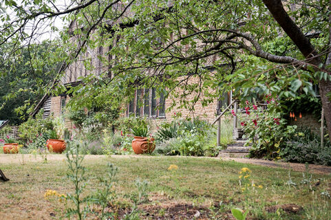 Photograph of the lawn outside Limnerslease: artists' home. The foliage interrupts the view of the fascia of the house, and there are three terracotta planters placed in a row by the exterior of the building