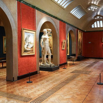 A wide-angle view inside a traditional art gallery. The walls are a rich, textured red, adorned with numerous framed oil paintings, many featuring golden frames. Down the centre of the gallery, a light-coloured marble statue of a female figure stands prominently between two arched alcoves. The floor is made of highly polished wooden parquet with dark decorative borders. Overhead, the ceiling is vaulted with skylights, providing natural light, supplemented by track lighting.