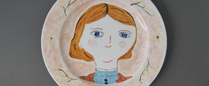 A white ceramic plate is centrally decorated with a stylized, whimsical portrait of a person. They have short, reddish-orange hair parted on the side, large blue eyes, small red lips, and pale skin. They wear a blue top with a black buttoned collar and a hint of an orange jacket. The rim of the plate is a soft peach colour with simple drawings of yellow flowers with blue centers and green stems. The plate is resting on a plain grey surface.