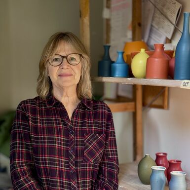 Lucy Burly with short blonde hair and glasses smiles directly at the camera while wearing a plaid shirt, positioned in front of a shelf displaying various colourful ceramic vases.