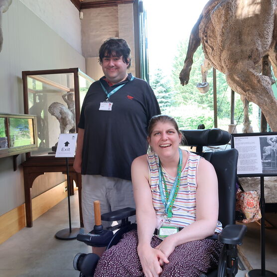 A women sitting in a wheelchair smiling wearing a striped vest and lanyard, with a man stood next to her wearing a black t-shirt