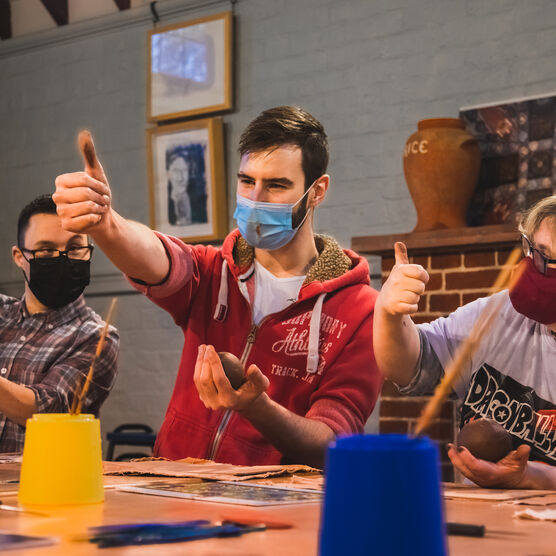 A photo of three people sat at a table working with clay wearing masks putting their thumbs up