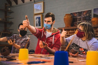 A photo of three people sat at a table working with clay wearing masks putting their thumbs up