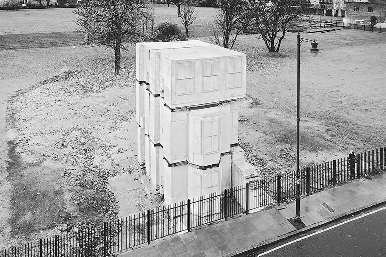 Black and white photo of concrete blocks in the abstract shape of a detached terrace house standing at the edge of a field by the pavement