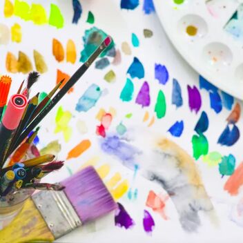 A vibrant image of an artist's workstation with paintbrushes in a jar, a palette with various colours, and scattered colourful paint swatches across a white surface.