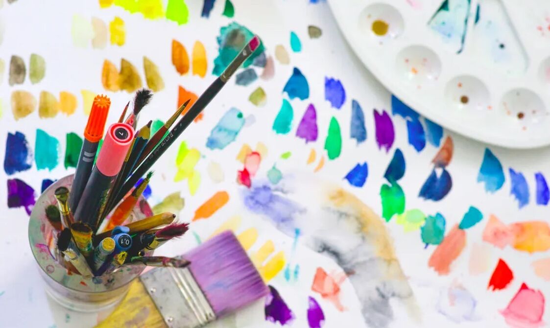 A vibrant image of an artist's workstation with paintbrushes in a jar, a palette with various colours, and scattered colourful paint swatches across a white surface.