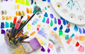 A vibrant image of an artist's workstation with paintbrushes in a jar, a palette with various colours, and scattered colourful paint swatches across a white surface.