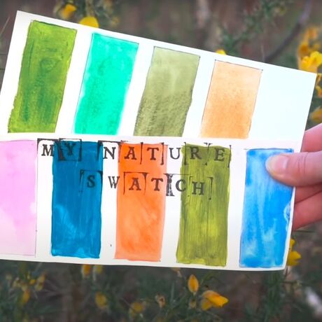 A hand holding a swatch book titled "my nature swatch" featuring various watercolour patches in shades of green, orange, pink, and blue, against a background of yellow flowers.