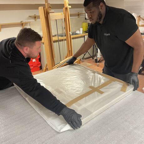 Two men carefully wrapping an oil painting in tissue and plastic