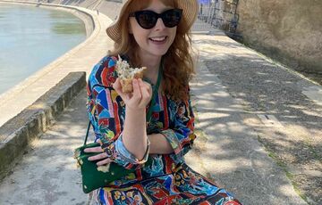 A woman with long red hair, in a wide-brimmed hat, sunglasses and a colourful dress eats bread by a river bank.