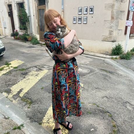 A woman with long red hair and a colourful dress holds a grey and white cat in a street in Arles.