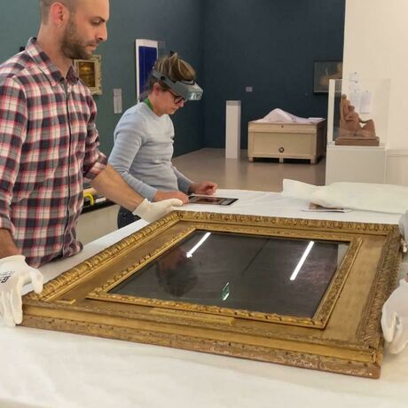 Two men in white gloves carefully place the painting on a table covered with tissue. A conservator consults her notes in the background.