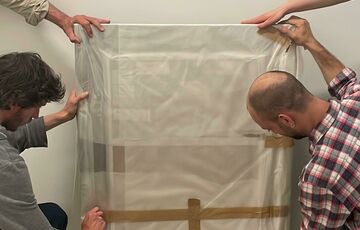 Art handlers carefully unwrap a painting wrapped in plastic sheeting and tissue. Two hands are holding the painting steady.