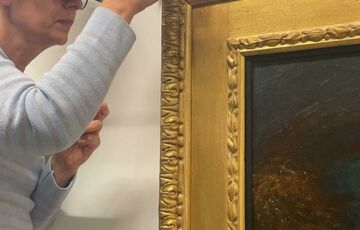A woman with magnifying goggles repairs a section of a gilt wooden frame