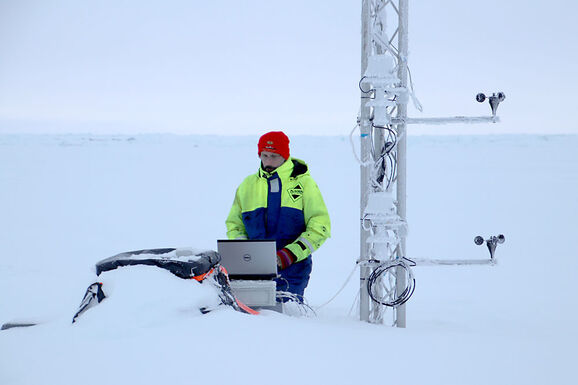 Photo of a man in the North Pole sitting with a computer by a metal mast in the snow. He is wearing a red hat and high-vis coat