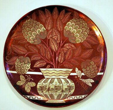 Red lustreware dish featuring a gold and cream coloured vase with dahlias in. There are two bees around the vase