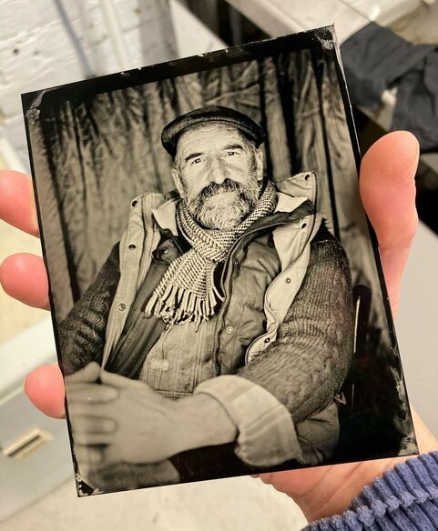 Tintype of a man in his 60s wearing a flat cap and lots of layers on looking in the camera. He has a beard.