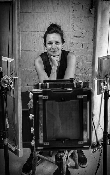 Photo of a woman smiling at the camera with a large format camera in the foreground in front of her.