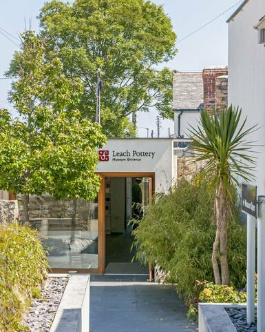 Photo of the entrance to Leach Pottery in St Ives. The building is white and the door is in the middle of the image with a shrub and palm tree in front of it to the right.
