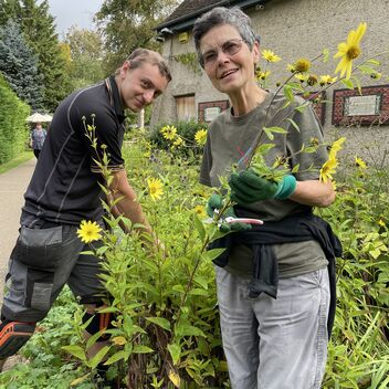 Photo of a short, grey-haired woman wearing khaki coloured clothes, gardening whilst smiling at the camera. There is a young man to the left of her helping