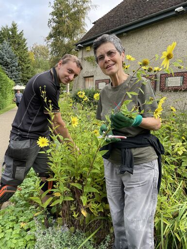 Photo of a short, grey-haired woman wearing khaki coloured clothes, gardening whilst smiling at the camera. There is a young man to the left of her helping