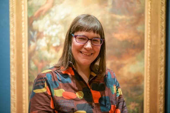 Laura wears a colourful shirt and stands in front of a painting