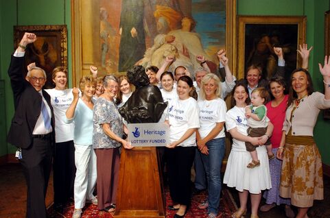 A group of smiling volunteers celebrate, they hold a plaque that read 'Heritage Lottery funded'