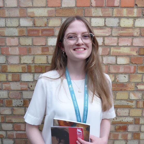A young, blonde women stands smiling for the camera in front of a brick wall, she holds a Historic Gallery guide booklet