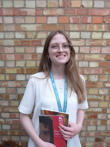 A young, blonde women stands smiling for the camera in front of a brick wall, she holds a Historic Gallery guide booklet
