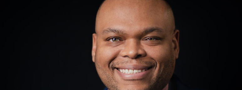 A photograph of a man looking at the camera smiling with short black hair wearing a blue suit, pink shirt and grey jumper
