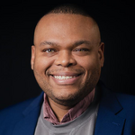 A photograph of a man looking at the camera smiling with short black hair wearing a blue suit, pink shirt and grey jumper