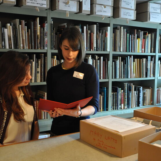 Two members of staff look at a book in the archive, there is a shelf of books behind them