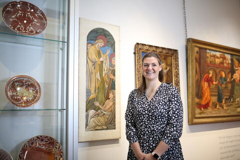 A women stands in the gallery with paintings and ceramics behind her