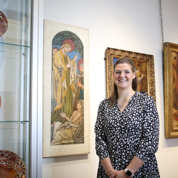 A women stands in the gallery with paintings and ceramics behind her
