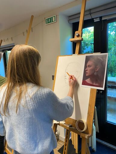 A woman with blonde hair in a blue jumper works on a sketch of another woman's silhouette at an easel