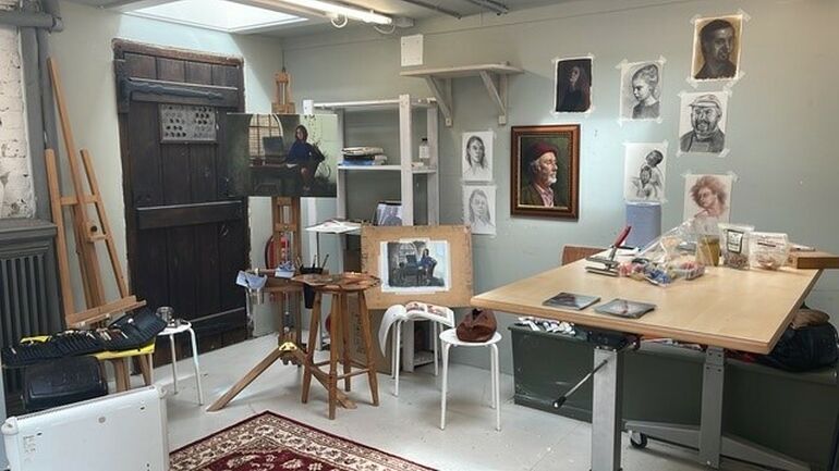 An artist's studio containing an easel and several stools, and a table covered in art supplies. On the wall are several portraits and pictures of G F Watts work