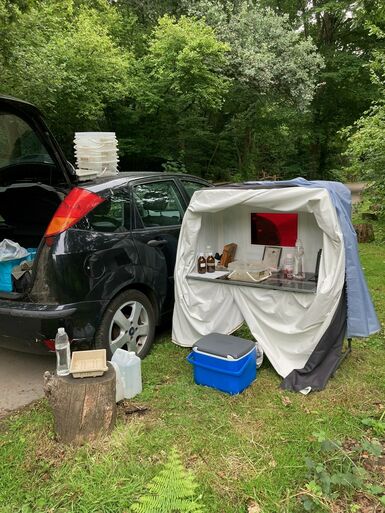 Photograph of the back of a black Ford car with the boot open. Next to it is a portable darkroom.
