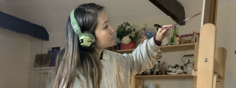 Sarah wears headphones and works on a painting on an easel