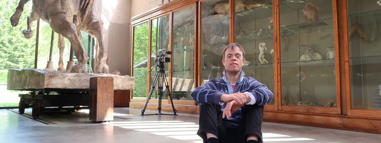 Chris sits on the concrete floor in the Sculpture Gallery, the large sculpture Physical Energy and a camera can be seen behind him