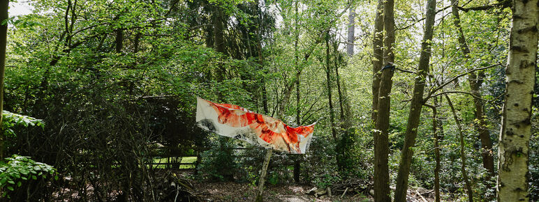 Photo of an abstract sculpture suspended in between trees in a woodland, made from canvas, featuring expressive orange paint marks