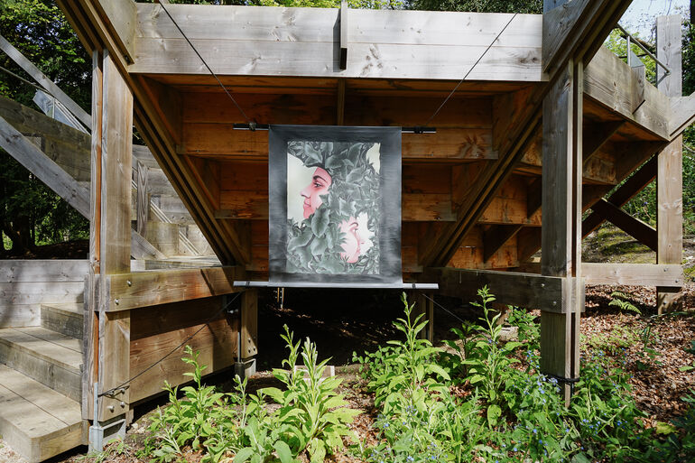Photo of Bench 1 in the Limnerslease woodland at Watts Gallery with a painting displayed on the side of two side profile faces