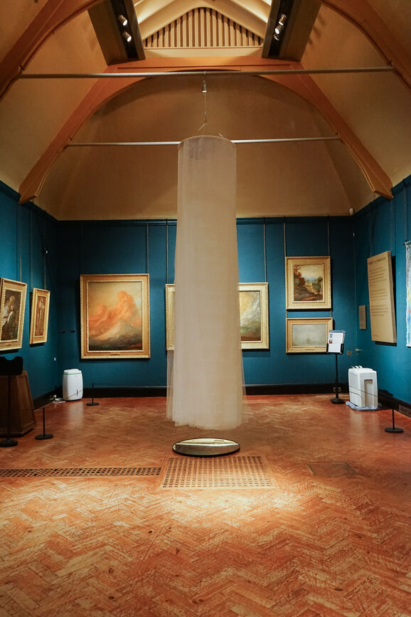 Photo inside the Historic Galleries at Watts Gallery. In the middle of the photo is an installation hanging from the ceiling made from tulle with a mirror underneath