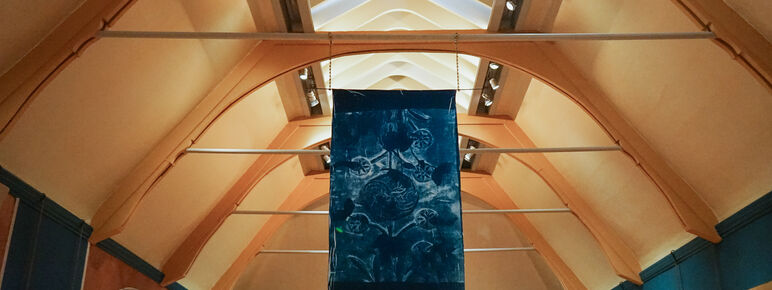 Cyanotype hanging from the ceiling of Watts Gallery with beading decorating the bottom of the fabric. There is an abstract design on the fabric